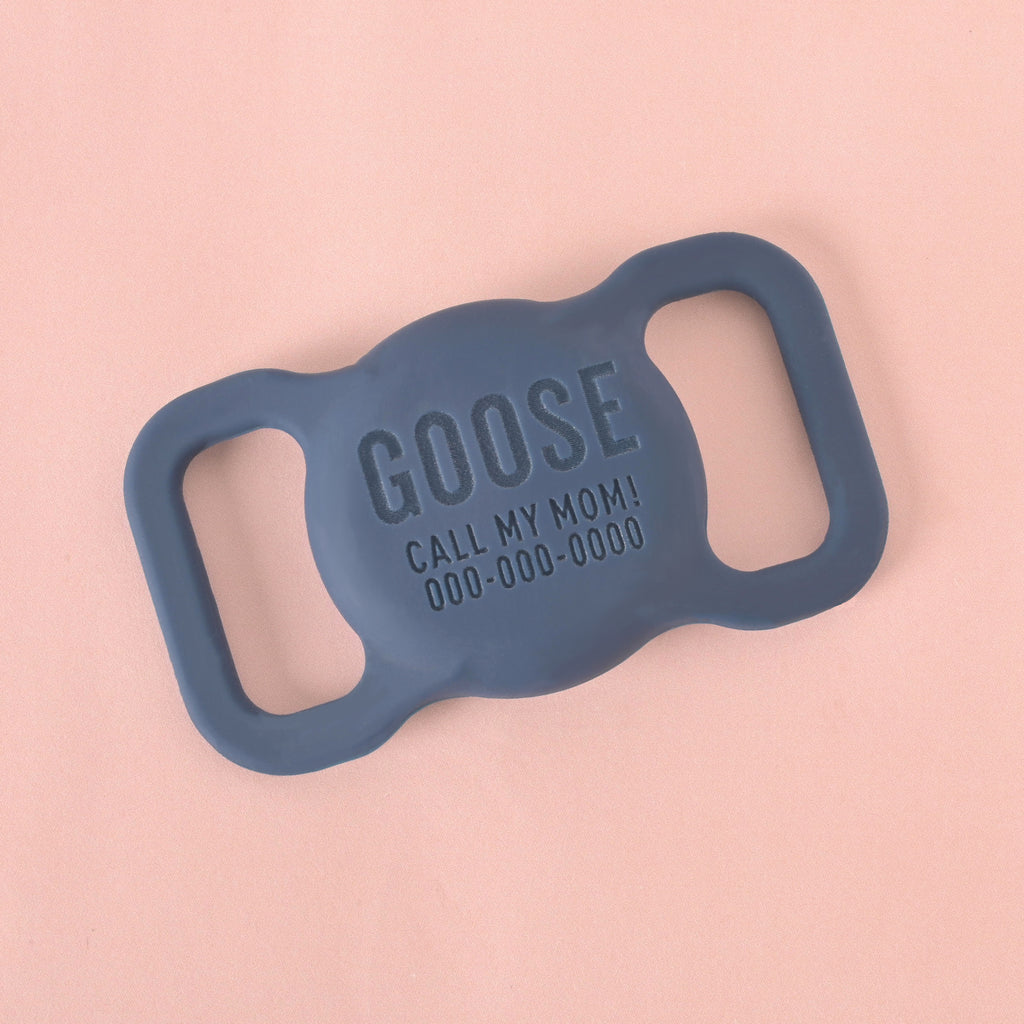 A close-up shot of a custom, silent pet ID tag designed to slide onto a collar. The tag is a navy blue color and is engraved with the name "GOOSE," the phrase "CALL MY MOM!", and a phone number below it. It is displayed on a solid pink background.