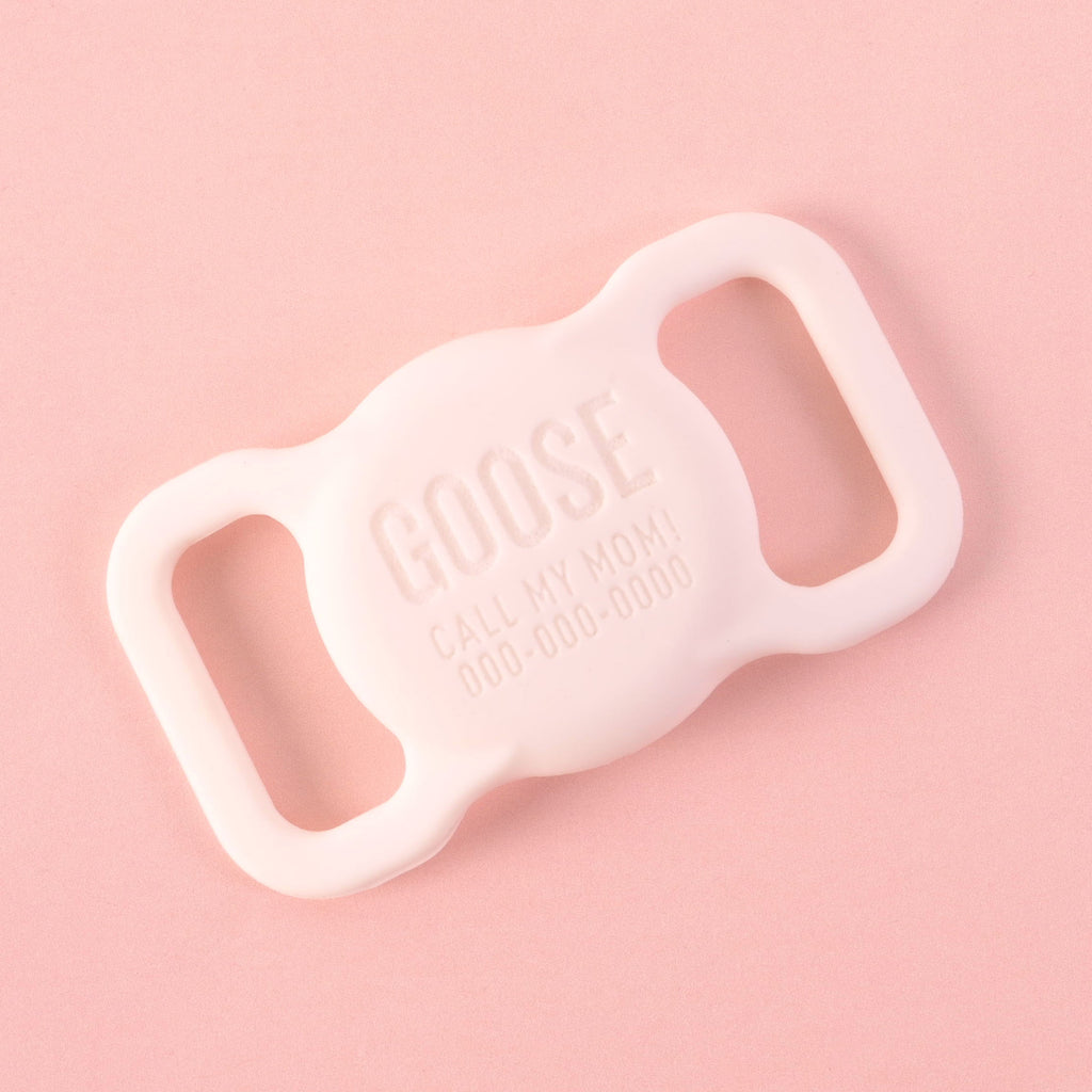 A personalized white AirTag holder for pets that says "GOOSE" and "CALL MY MOM!" with a placeholder phone number on it. The holder is displayed on a solid pink background.