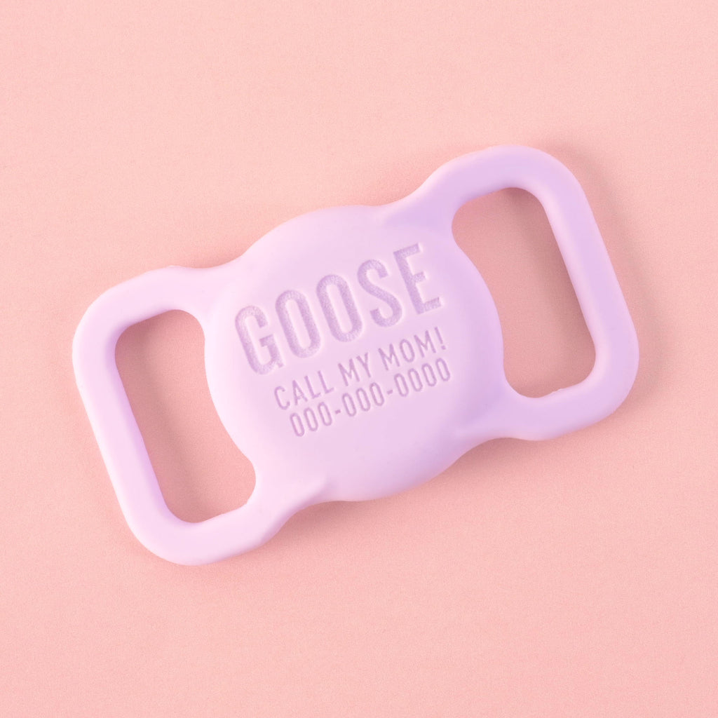 A personalized lavender AirTag holder for pets that says "GOOSE" and "CALL MY MOM!" with a placeholder phone number on it. The holder is displayed on a solid pink background.