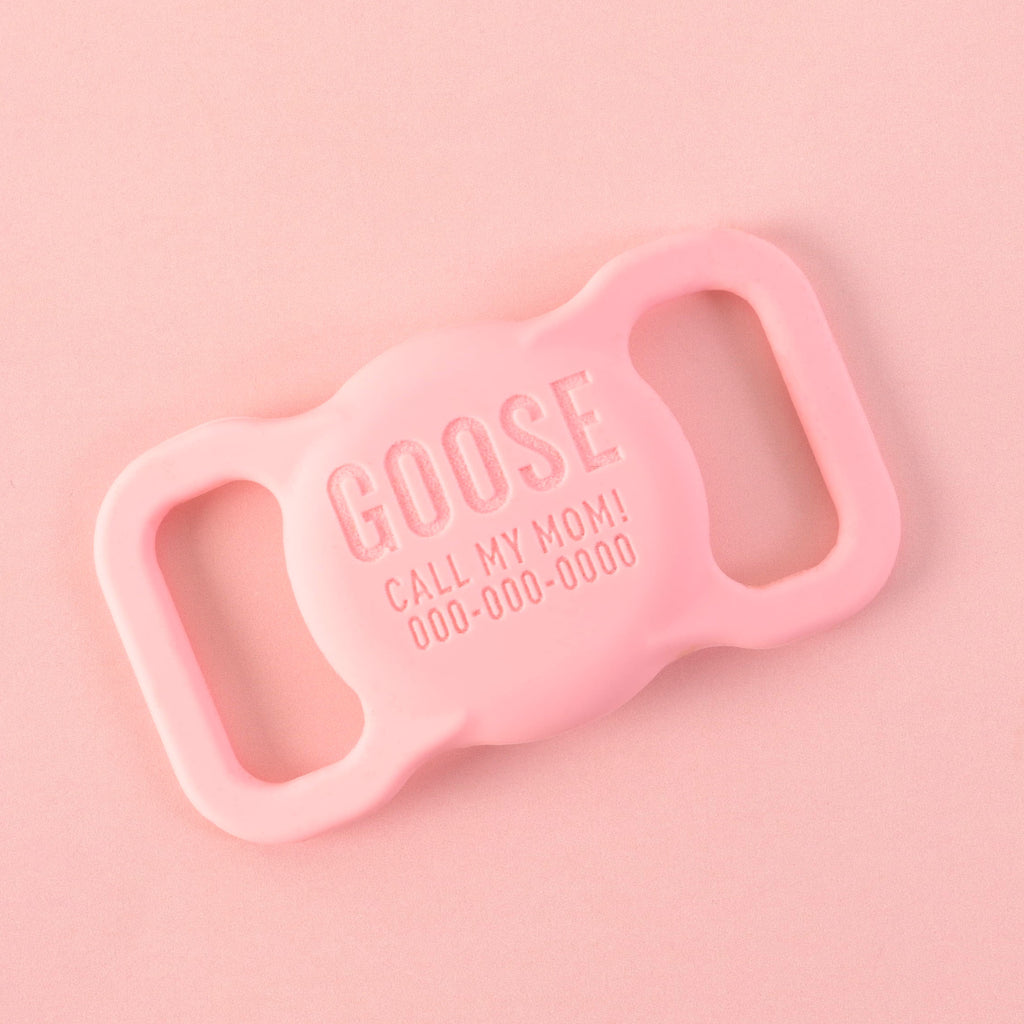 A personalized pink AirTag holder for pets that says "GOOSE" and "CALL MY MOM!" with a placeholder phone number on it. The holder is displayed on a solid pink background.