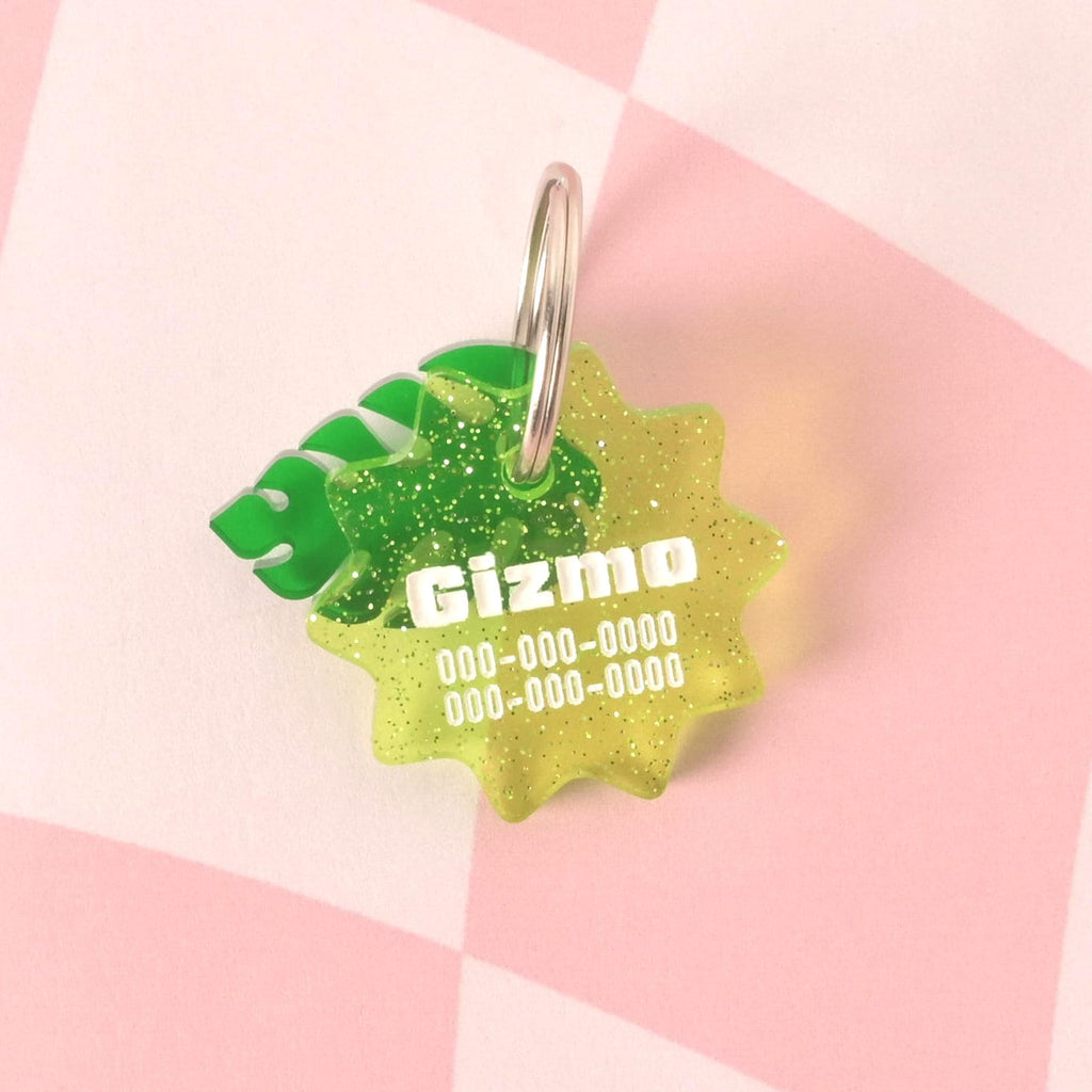 A close-up of a custom, glittery green, starburst-shaped pet ID tag. The tag is engraved with the name "Gizmo" and a phone number in white font. Two green tropical leaf charms are attached to the silver keyring. The tag is displayed on a pink and white checkered background.