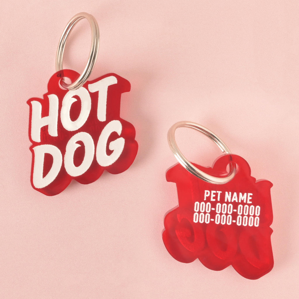 A red pet tag in the shape of a speech bubble. The front says "HOT DOG" in a white groovy font, and the back shows a placeholder for the pet's name and phone number. The tags are resting on a solid pink background.