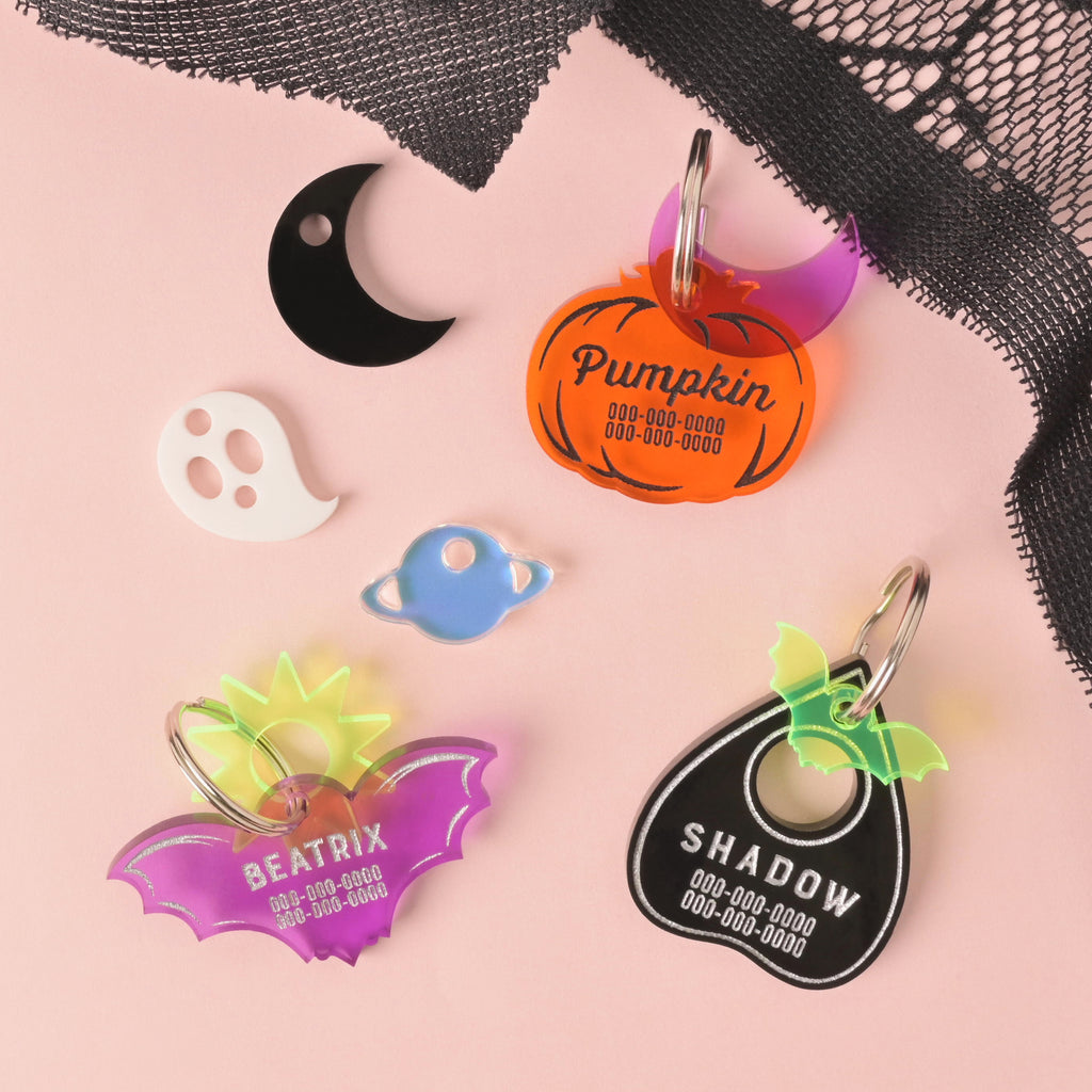 A collection of custom acrylic pet tags and mini charms: a personalized orange pumpkin tag, a purple bat tag, a black planchette tag with a neon bat accent, a black crescent moon charm, a white ghost charm, and a blue planet charm, displayed on a pink background with black lace.