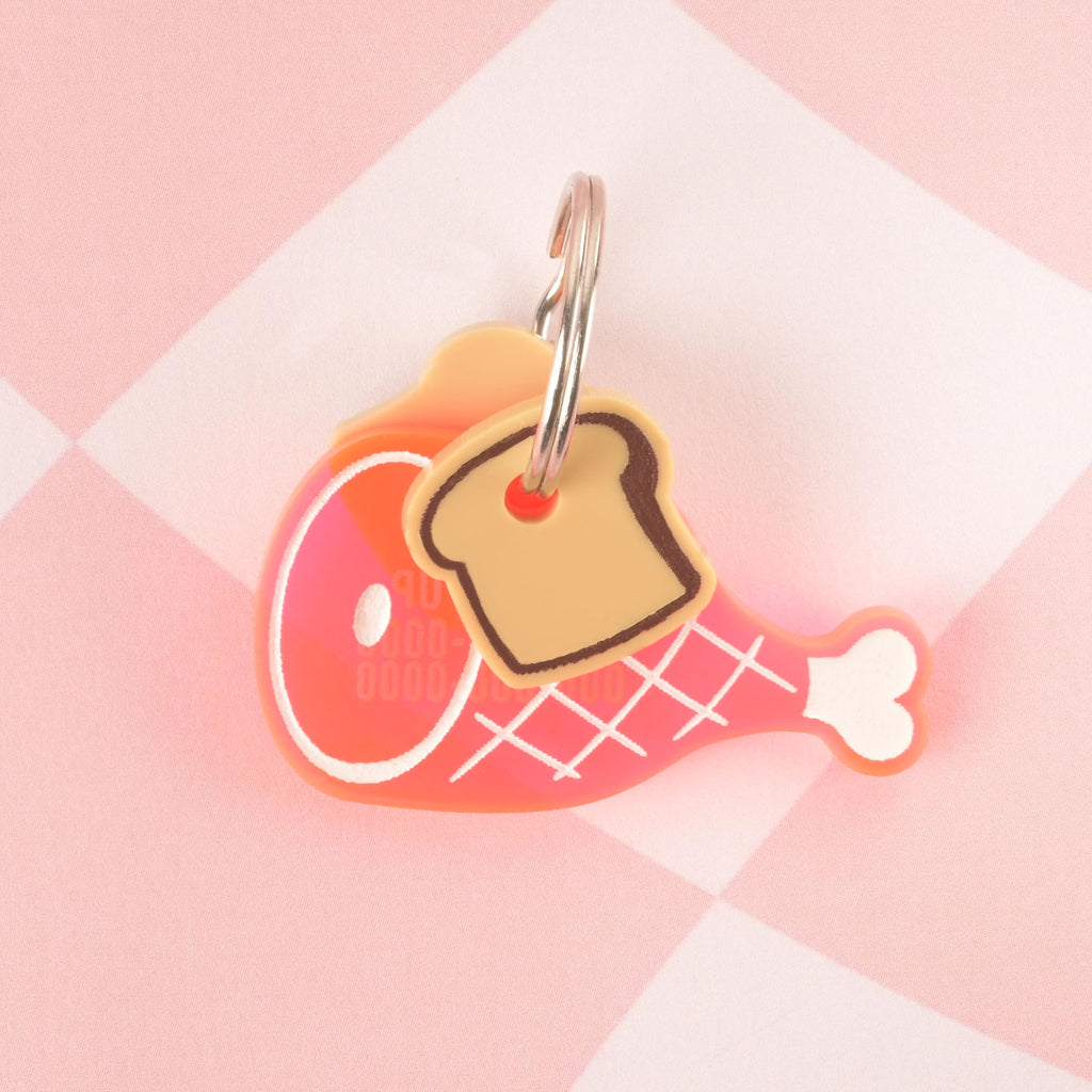 A large pink ham-shaped pet tag with white grid and bone details. A small, tan toast-shaped charm is attached to the tag's ring. The tag has a hole for the attached silver ring and is resting on a pink and white checkered background.