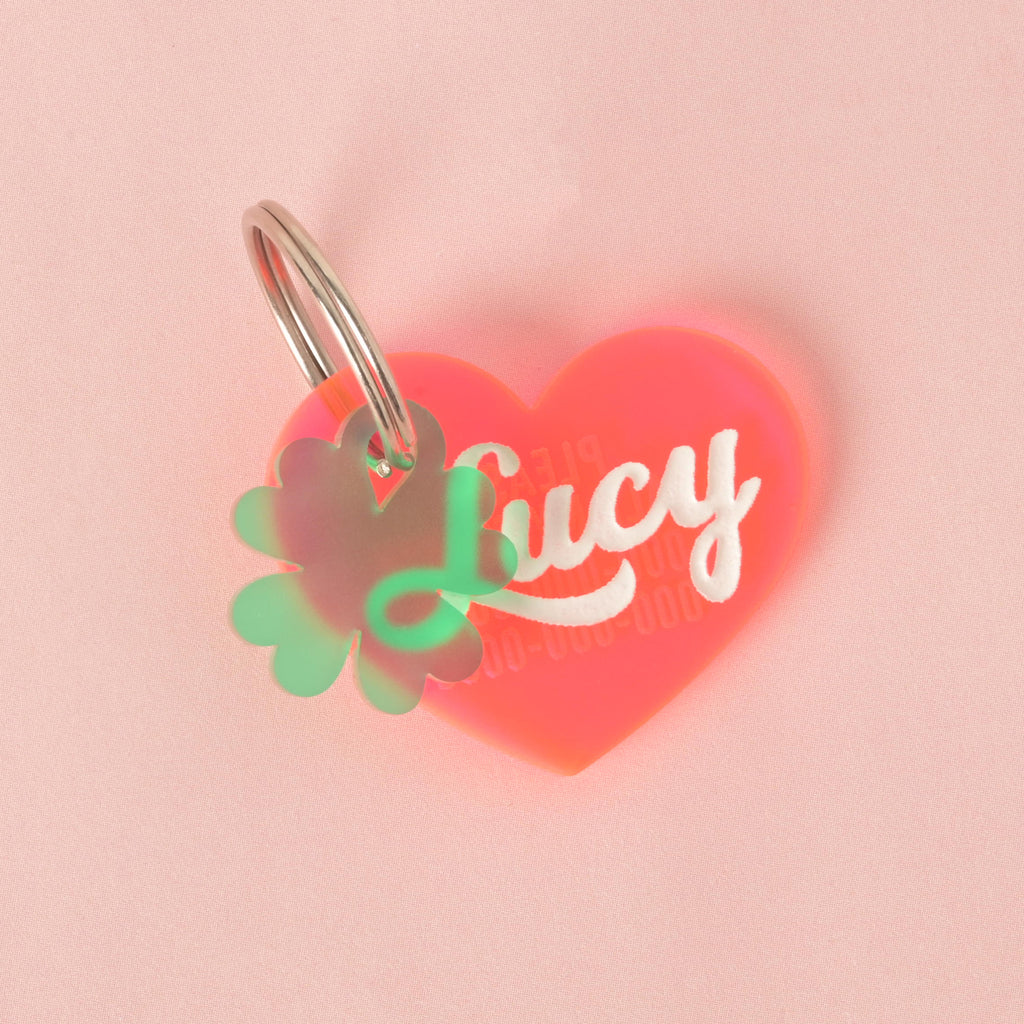 A custom, translucent orange heart-shaped pet ID tag. The tag is engraved with the name "Lucy" in white script font and a phone number below it. A second translucent green, four-leaf clover charm is attached, sitting in front of the main tag. Both are on a silver keyring and are displayed on a solid pink background.