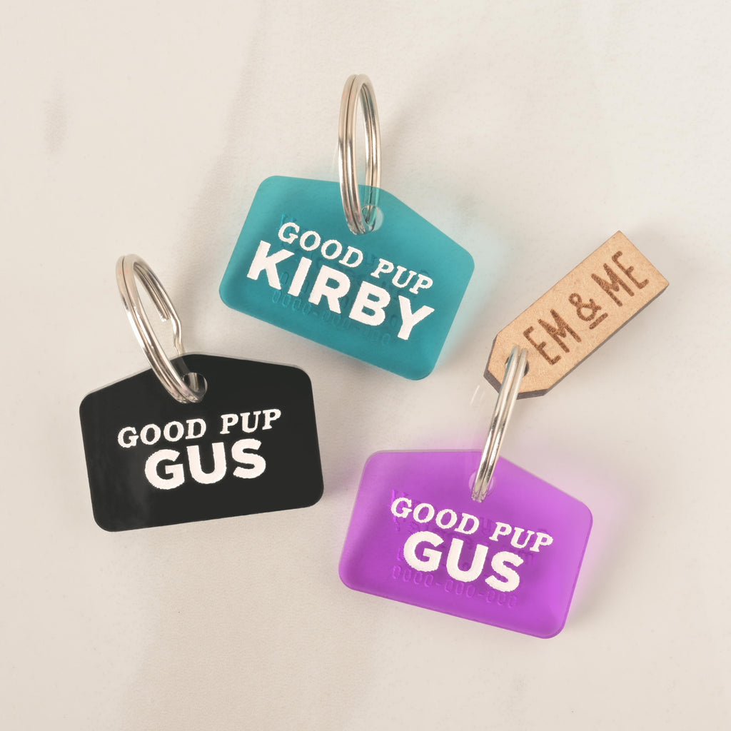 A set of three "Good Pup" dog tags in different colors. One black tag says "Gus," a blue-green tag says "Kirby," and a purple tag says "Gus." All tags have a placeholder phone number and are attached to a silver ring for a collar. A small wooden charm that says "EM & ME" is also visible. The items are shown on a light-colored surface.