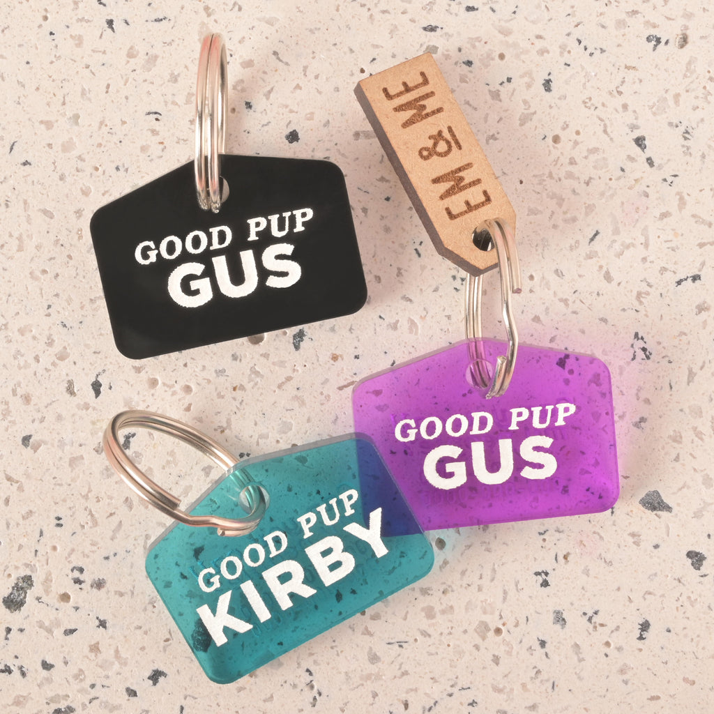 A set of three "Good Pup" dog tags in different colors. One black tag says "Gus," a blue-green tag says "Kirby," and a purple tag says "Gus." All tags have a placeholder phone number and are attached to a silver ring for a collar. A small wooden charm that says "EM & ME" is also visible. The items are shown on a light-colored, speckled surface.