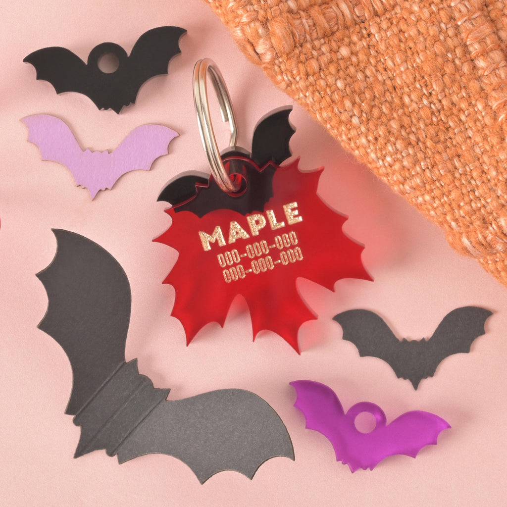 A personalized red maple leaf-shaped pet tag with the name "MAPLE" written in gold letters, along with a placeholder phone number. The tag has a small, black bat charm attached behind it. The tags are connected to a silver ring for a collar and are displayed on a solid pink background with several other bat-shaped charms in black, gray, and purple. A brown knitted blanket is in the top right corner.