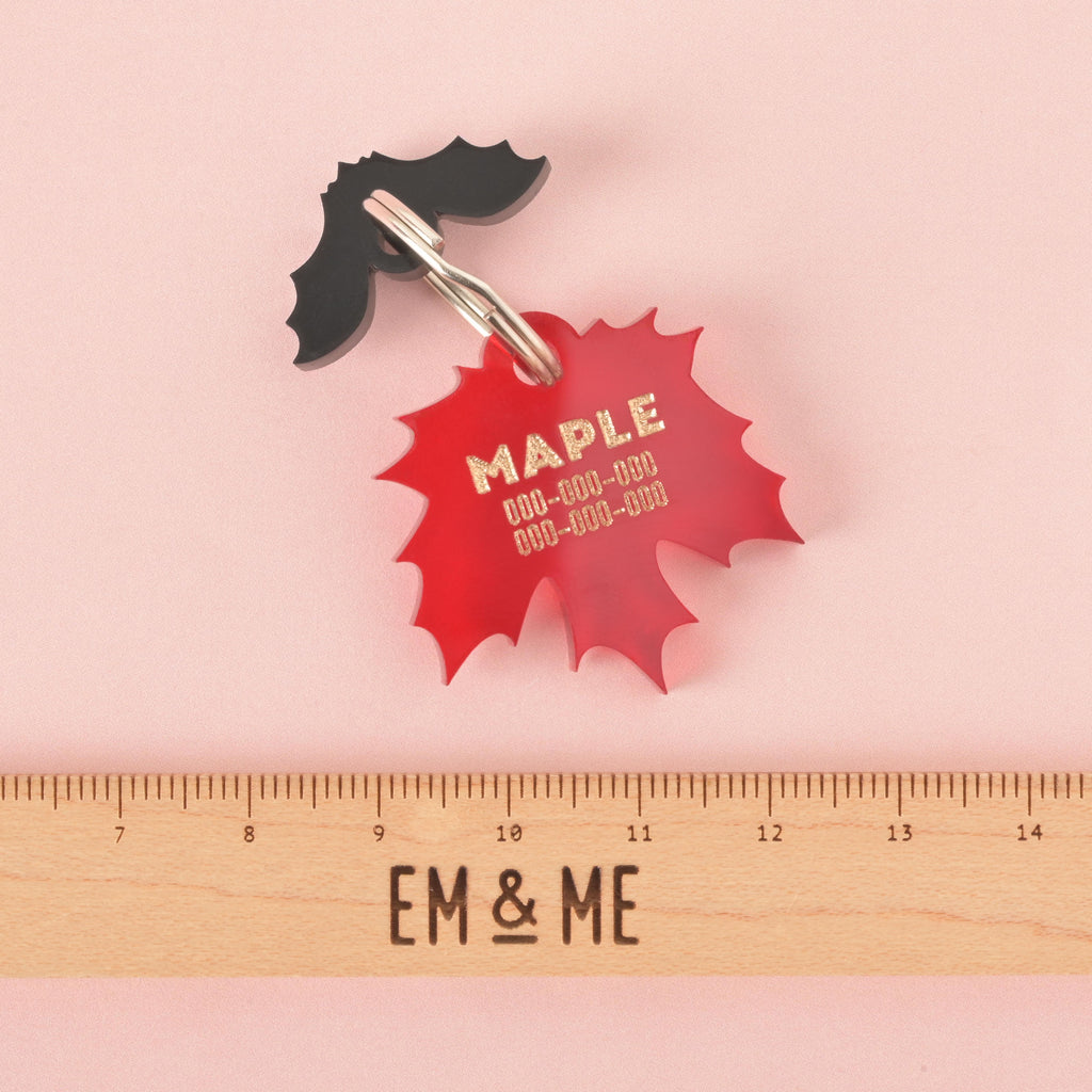 A personalized red maple leaf-shaped pet tag with the name "MAPLE" written in gold letters, along with a placeholder phone number. The tag has a small, black bat charm attached behind it. The tags are connected to a silver ring for a collar and are displayed on a solid pink background.
