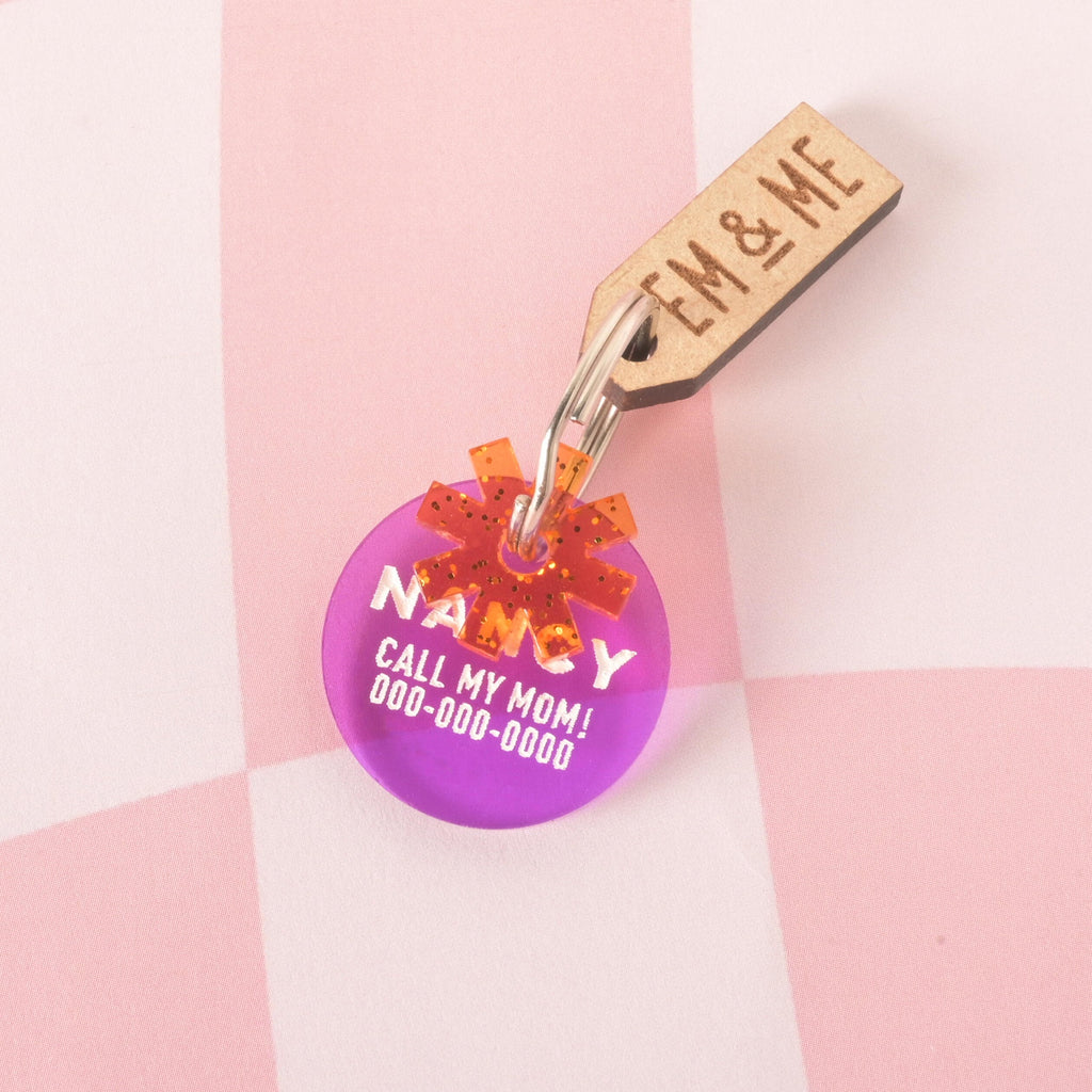 Flower charm, Orange Asterisk Mini, Geometric Flower Seasonal Mini, Tiny Accessory, Add-on Charm A custom, translucent purple, circular pet ID tag. The tag is engraved with the name "NANCY," the phrase "CALL MY MOM!", and a phone number in white font. A small, glittery orange starburst charm is attached, sitting in front of the main tag. A wooden tag with the logo "EM & ME" is also visible. The tags are attached to a silver keyring and are displayed on a pink and white checkered background.