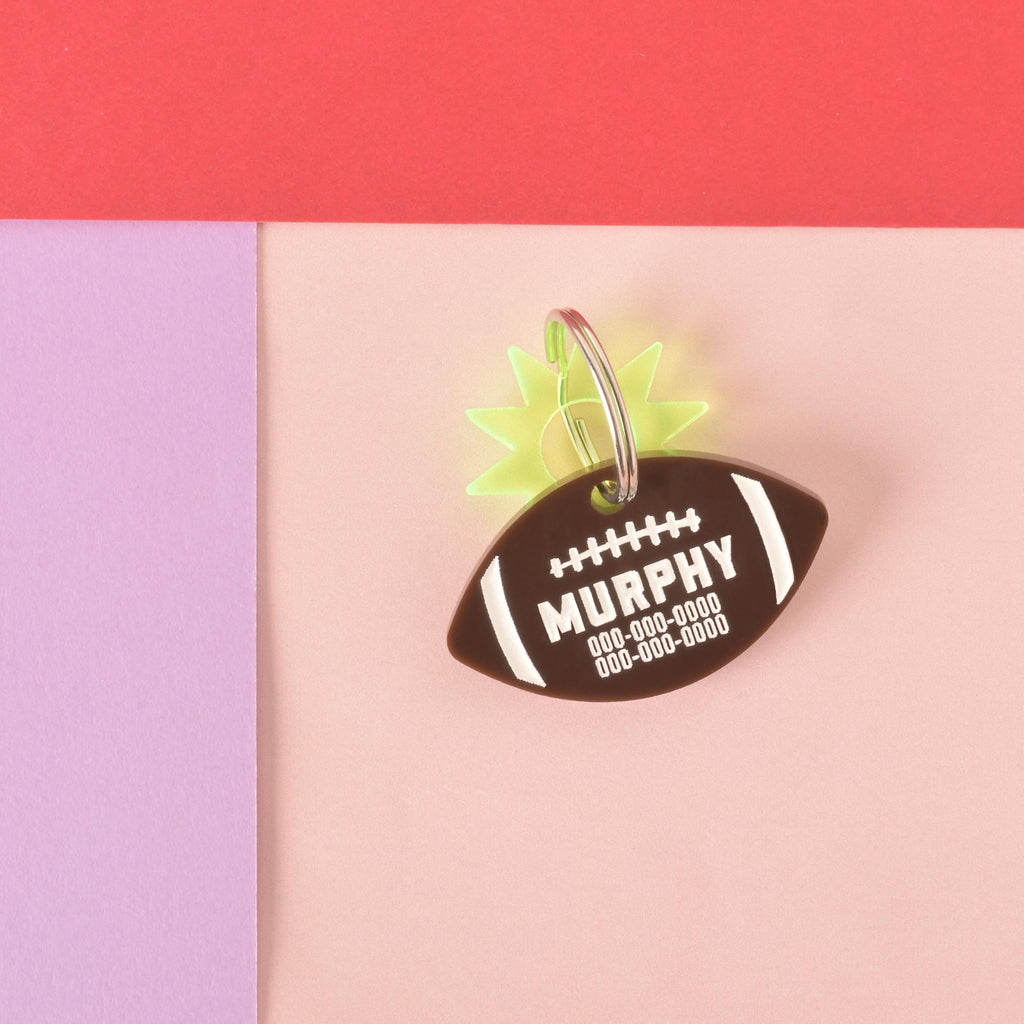 A personalized brown football-shaped pet tag with the name "MURPHY" and a placeholder phone number on it. A translucent yellow starburst charm is attached behind the tag. Both are connected to a silver ring, perfect for a collar, and are displayed on a light pink, purple, and red background.
