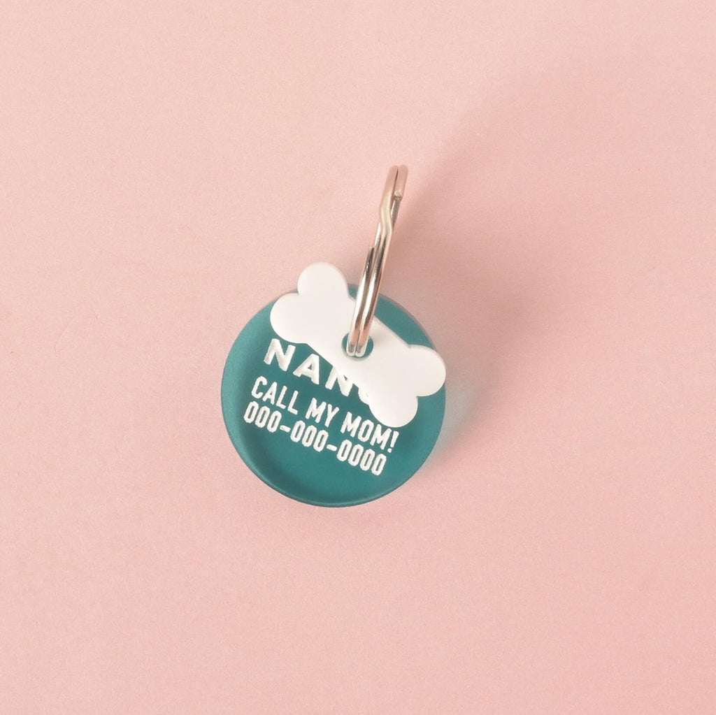 A close-up shot of a custom, round, translucent teal pet ID tag. The tag is engraved with the name "NANCY," the phrase "CALL MY MOM!", and a phone number in white font. A small white bone charm is attached, sitting on top of the tag. Both are on a shiny silver keyring and are displayed on a solid pink background.
