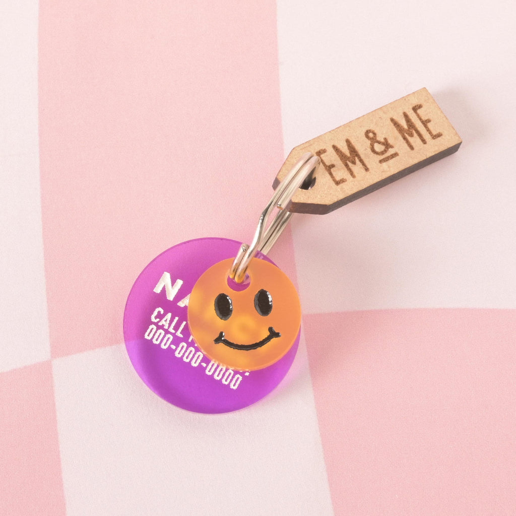 A custom, purple, circular pet ID tag. The tag is engraved with a name and a phone number. A small, orange smiley face charm is attached, sitting in front of the main tag. A wooden tag with the logo "EM & ME" is also visible. The tags are attached to a silver keyring and are displayed on a pink and white checkered background.