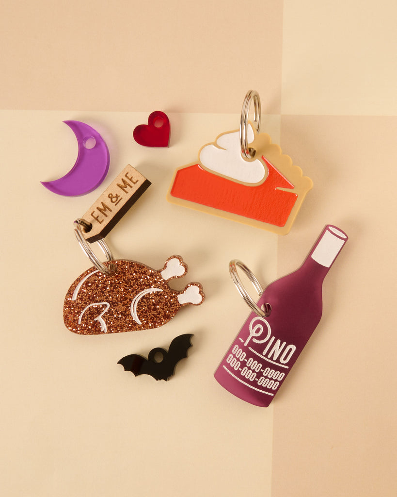A collection of custom pet tags and mini charms: a layered pie slice tag with whipped cream, a glitter brown turkey/roast tag, a personalized wine bottle tag, a purple crescent moon, a red heart, a black bat, and a small wooden branded tag.