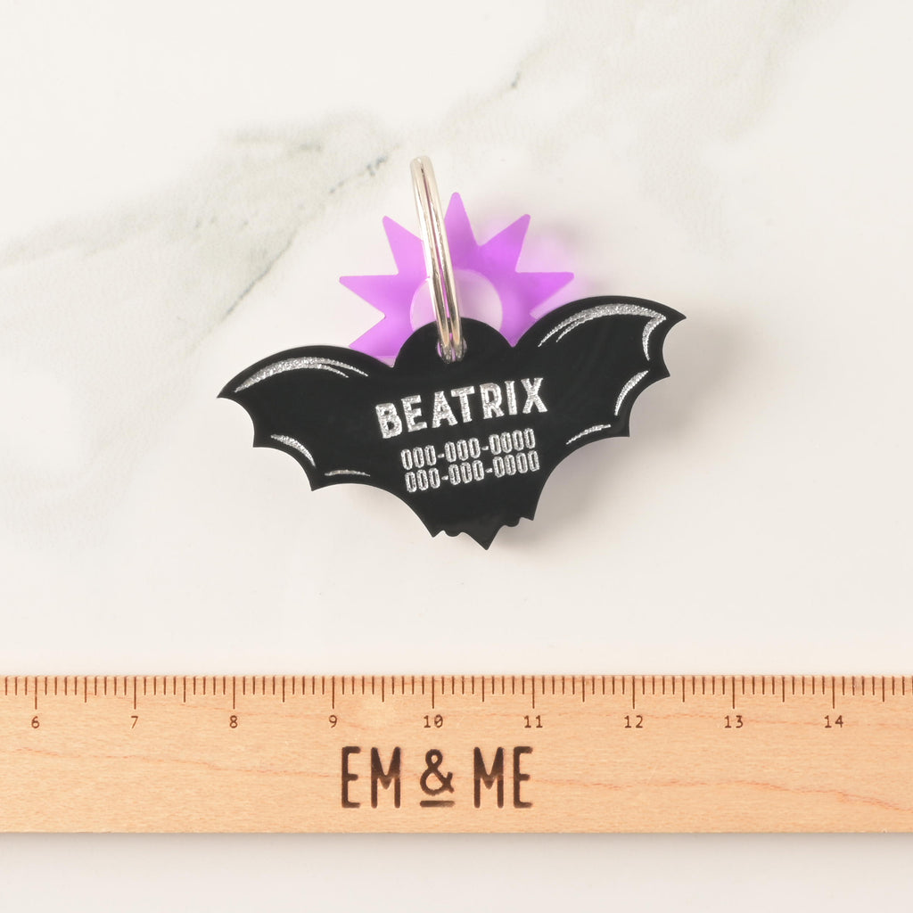 An opaque black acrylic bat-shaped pet ID tag, personalized with the name 'BEATRIX' and phone numbers. A translucent purple sunburst charm is attached to the ring. The tag is shown above a wooden ruler marked in centimeters to clearly indicate its size.