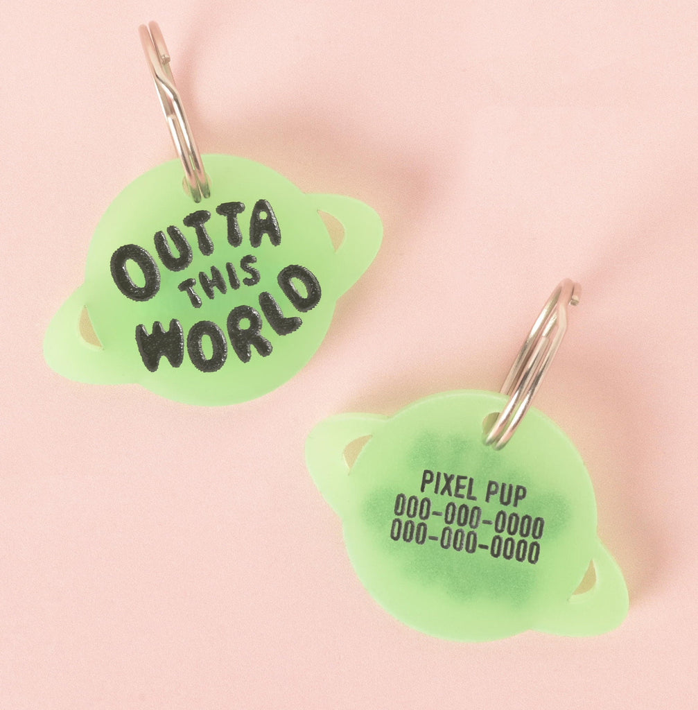 A pair of custom, glow-in-the-dark, planet-shaped pet ID tags. The tag on the left is engraved with "OUTTA THIS WORLD" in black font. The tag on the right is engraved with name and a phone number. Both are attached to silver keyrings and are displayed on a solid pink background.