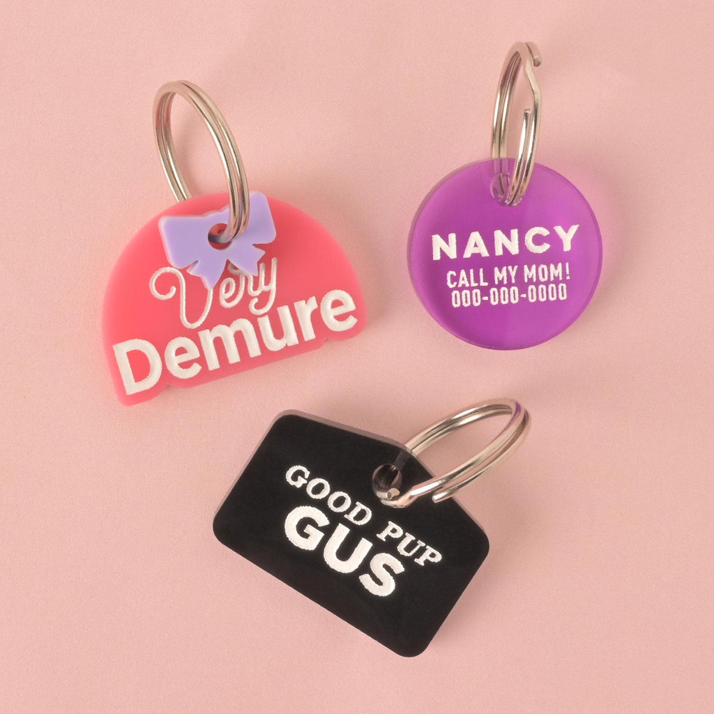  group of three personalized pet ID tags on a pink background. The tags are attached to silver keyrings. The tag on the top left is pink, half-moon shaped, with a light purple bow and the script words "Very Demure." The top right tag is a purple circle engraved with the name "NANCY," "CALL MY MOM!", and a phone number. The bottom tag is a black rectangle with the text "GOOD PUP GUS" in white font.