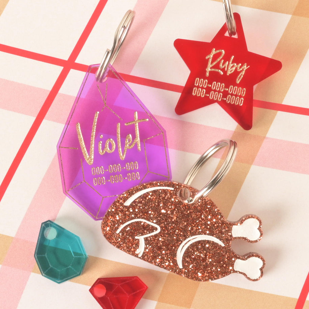 Acrylic pet ID tags including a glitter turkey shape, purple gemstone-shaped tag with engraved name Violet, and red star tag with engraved name Ruby, customizable with phone number for dogs and cats.