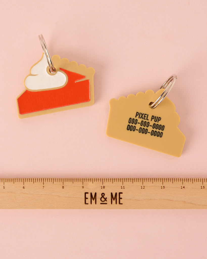 A personalized pet ID tag shaped like a slice of pumpkin pie, made from layered acrylic in tan, orange/red, and white whipped cream. The back is engraved with 'PIXEL PUP' and phone numbers. The tag is shown above a wooden ruler for size reference.