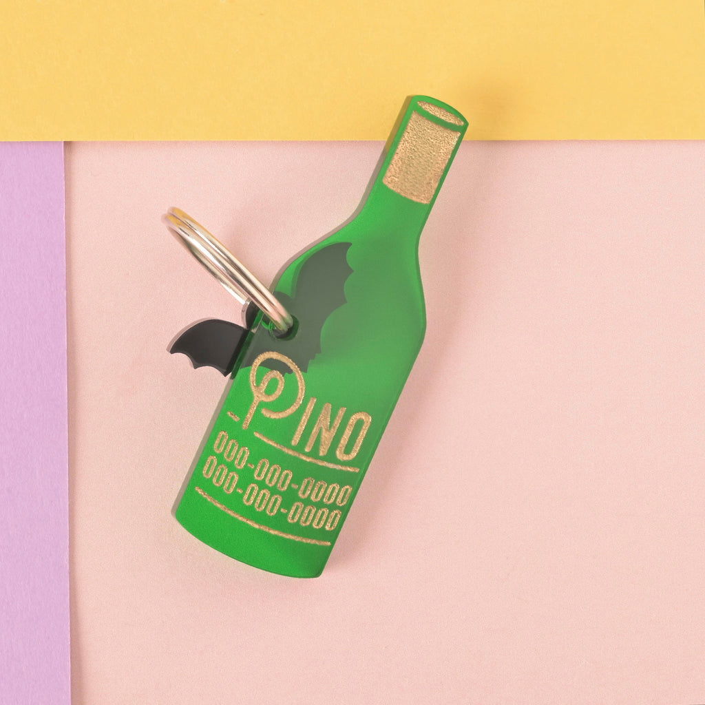 A personalized green wine bottle-shaped pet tag that says "Pino" in a gold-colored groovy font, along with a placeholder phone number. A small black bat charm is attached behind the tag. The tag is connected to a silver ring for a collar and is displayed on a light pink, purple, and yellow background.
