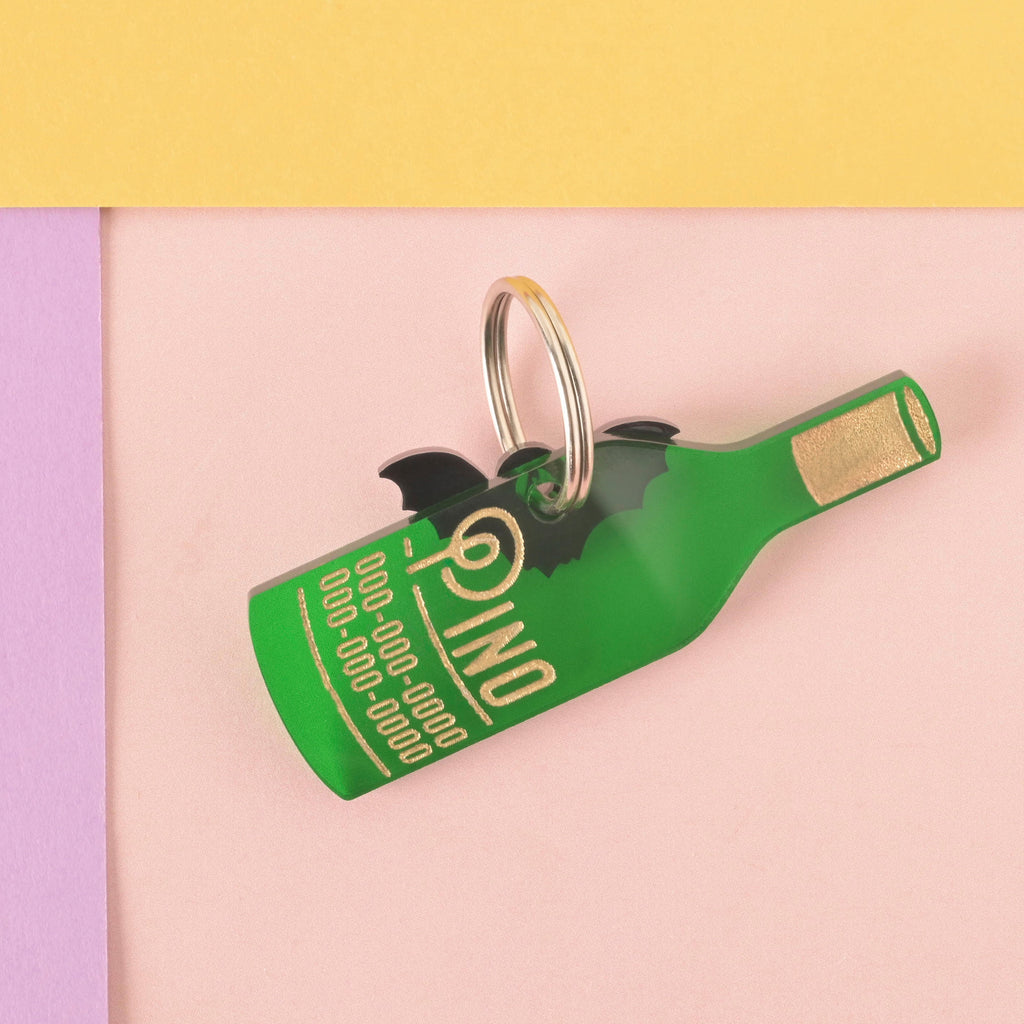 A personalized green wine bottle-shaped pet tag that says "Pino" in a gold-colored groovy font, along with a placeholder phone number. A small black bat charm is attached behind the tag. Both are connected to a silver ring for a collar and are displayed on a light pink and yellow background.