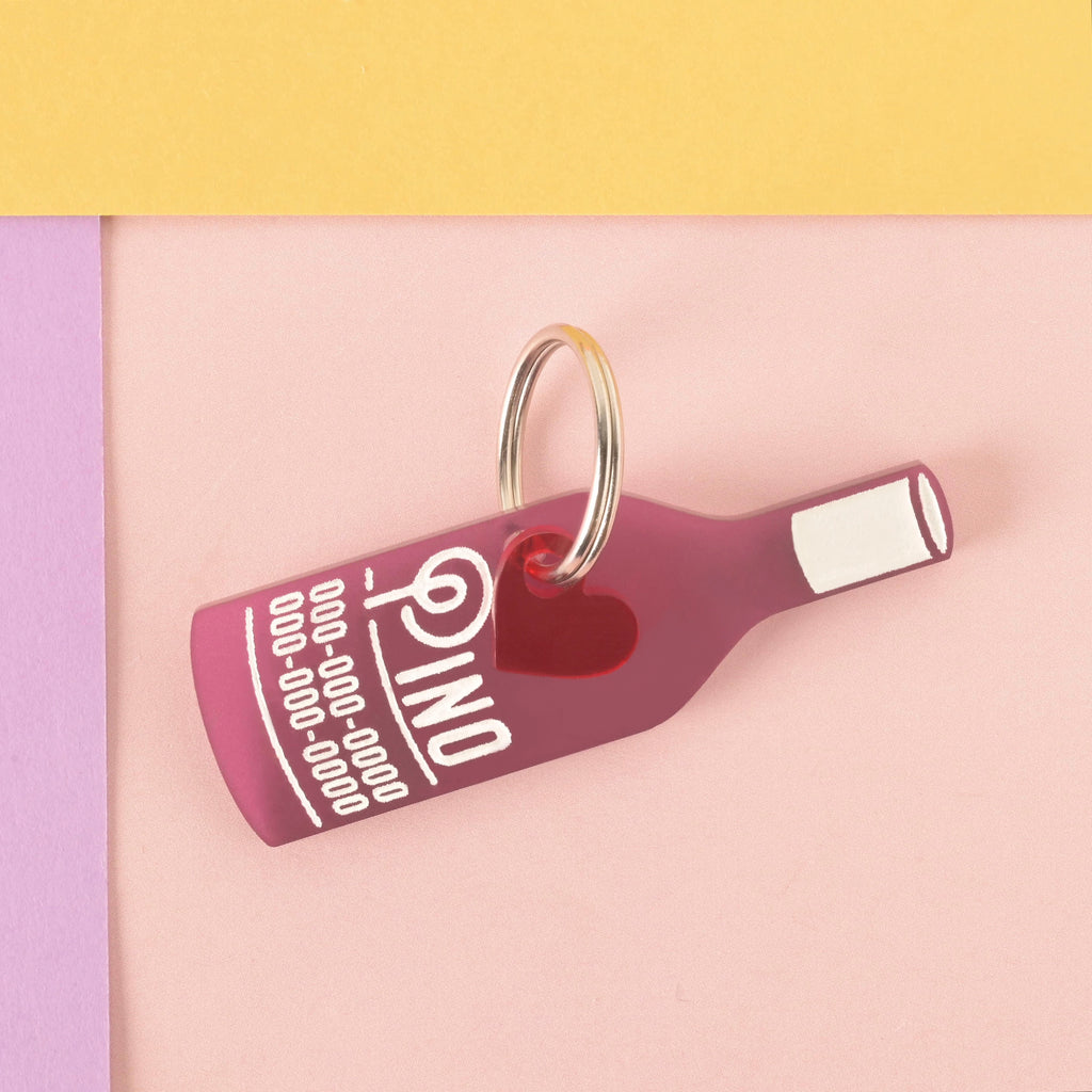 A personalized frosted wine-colored wine bottle-shaped pet tag that says "Pino" in a white groovy font, along with a placeholder phone number. A small red heart charm is attached behind the tag. The tag is connected to a silver ring for a collar and is displayed on a light pink, purple, and yellow background.