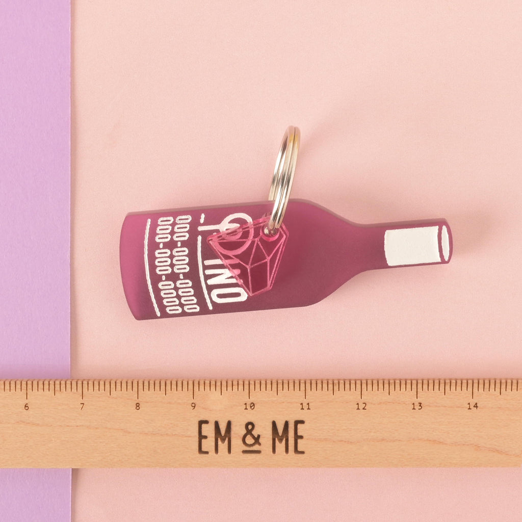 A personalized dark pink wine bottle-shaped pet tag that says "Pino" in a white groovy font, along with a placeholder phone number. A small pink diamond charm is attached behind the tag. The tags are connected to a silver ring for a collar. A wooden ruler with the "EM & ME" logo is placed below the tag to show its size. The items are displayed on a background of solid pink, purple, and red.