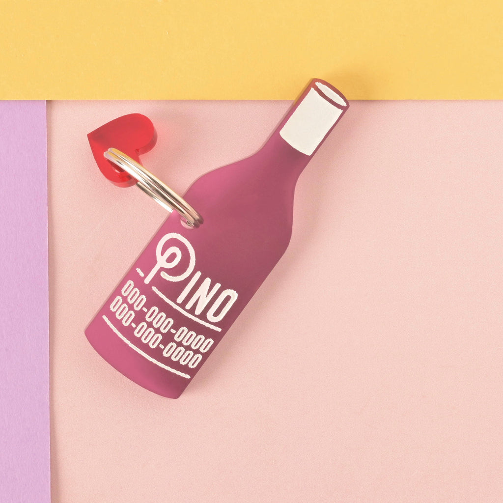A personalized dark pink wine bottle-shaped pet tag that says "Pino" in a white groovy font, along with a placeholder phone number. A small red heart charm is attached behind the tag. The tag is connected to a silver ring for a collar and is displayed on a light pink, purple, and yellow background.