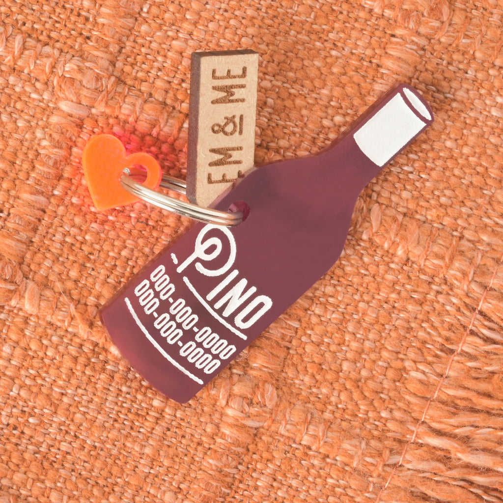 A personalized dark pink wine bottle-shaped pet tag that says "Pino" in a white groovy font, along with a placeholder phone number. The tag has a small, neon orange heart charm and a wooden "EM & ME" tag attached. The items are connected to a silver ring for a collar and are displayed on an orange knitted blanket.