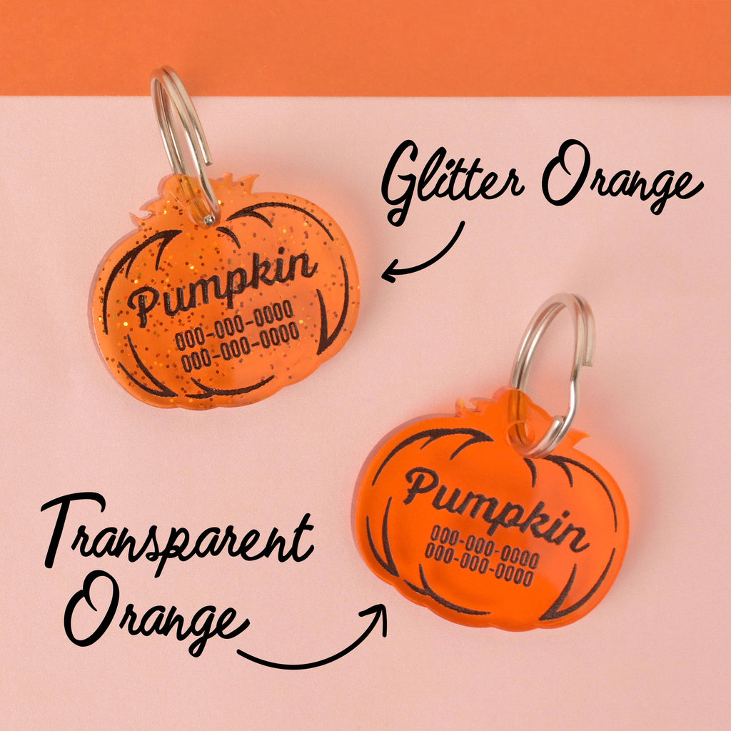 A comparison shot of two pumpkin-shaped pet ID tags. The top tag is labeled "Glitter Orange" and is a translucent tag with glitter specs, engraved with "Pumpkin" and a phone number. The bottom tag is labeled "Transparent Orange" and is a solid, translucent orange, also engraved with "Pumpkin" and a phone number. Both tags have silver keyrings and are displayed on a light pink and orange color-blocked background.