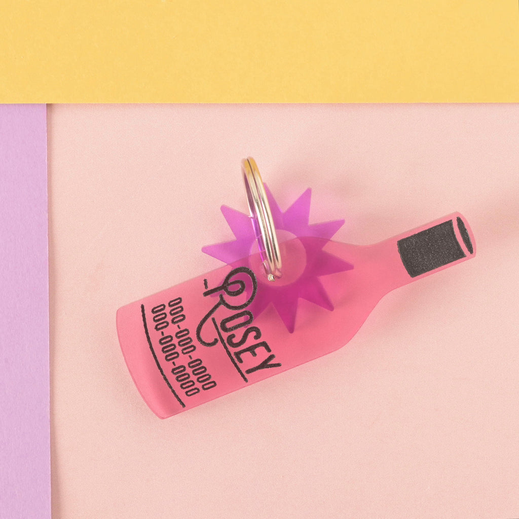 A personalized pink wine bottle-shaped pet tag with the name "ROSEY" and a placeholder phone number on it. The tag has a translucent purple starburst charm attached. The tag is connected to a silver ring for a collar and is displayed on a light pink, purple, and yellow background.
