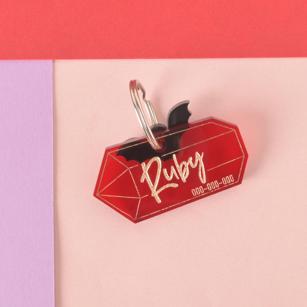 A personalized red gemstone-shaped pet tag with the name "Ruby" written in a gold-colored script, along with a placeholder phone number. A small black bat charm is attached behind the tag. Both are connected to a silver ring for a collar and are displayed on a light pink, purple, and red background.