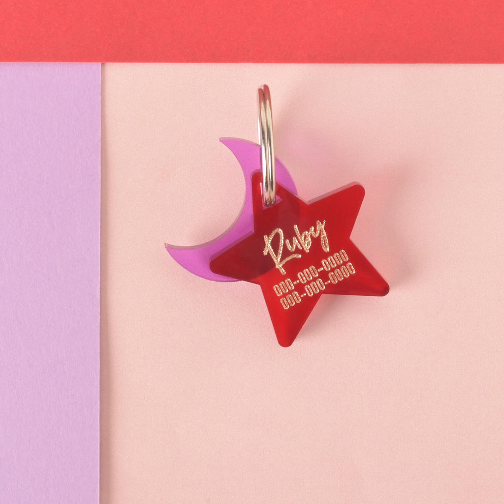 A personalized red star-shaped pet tag with the name "Ruby" and a placeholder phone number on it. A translucent purple crescent moon charm is attached behind the tag. The tags are connected to a silver ring for a collar and are displayed on a light pink, purple, and red background.