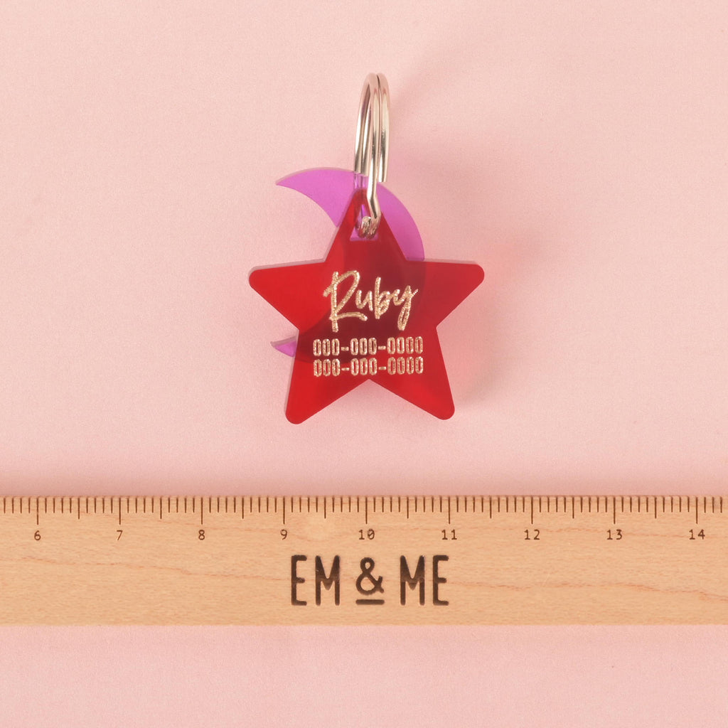 A personalized red star-shaped pet tag with the name "Ruby" and a placeholder phone number on it. A purple crescent moon charm is attached behind the tag. A wooden ruler with the "EM & ME" logo is placed below the tags to show their size. The items are displayed on a solid pink background.
