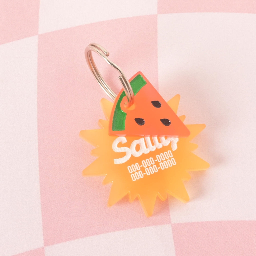 A custom, translucent orange, sun-shaped pet ID tag with a shiny silver keyring. The tag is engraved with the name "Sally" and a phone number in white font. A small, pink and green watermelon slice charm with black seeds is attached to the keyring, sitting in front of the main tag. The tags are displayed on a pink and white checkered background.