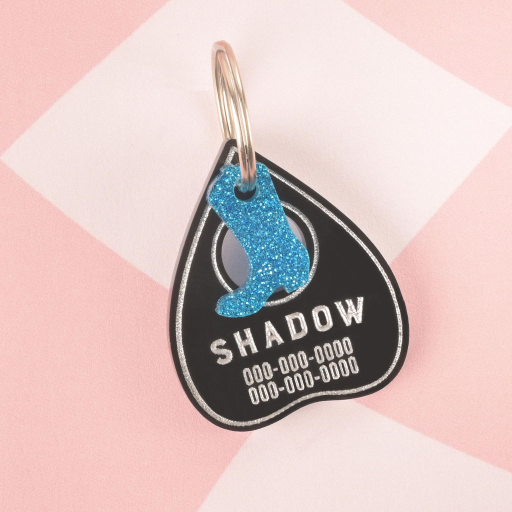 A personalized black Ouija planchette-shaped pet tag with the name "SHADOW" and a placeholder phone number. A blue glitter cowboy boot charm is attached to the tag's ring. Both are secured with a silver ring for a collar