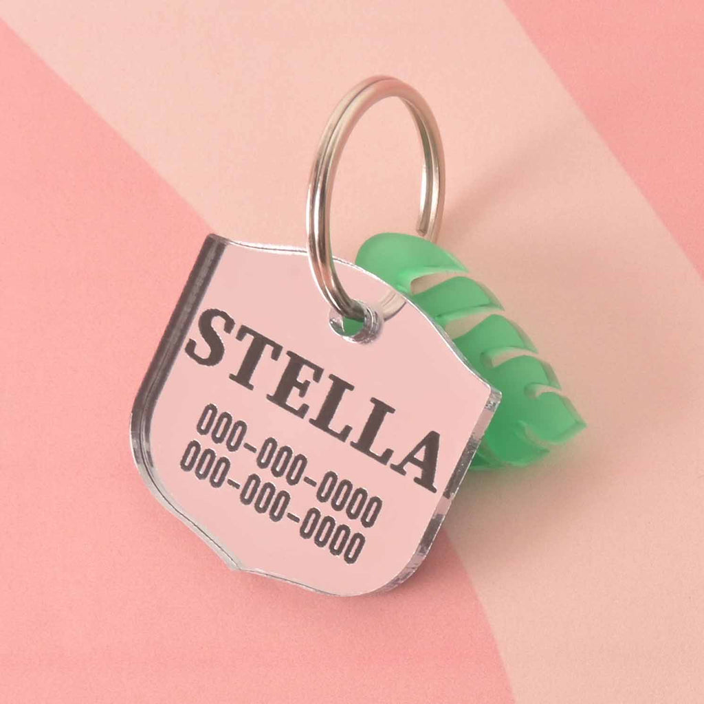 A personalized silver shield-shaped pet tag with the name "STELLA" and a placeholder phone number. The tag has a small, green monstera leaf charm attached. Both are connected to a silver ring, perfect for a collar, and are displayed on a light pink background with a wavy design.