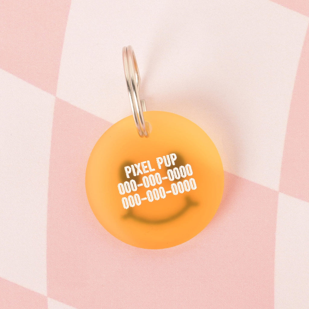 A custom, translucent orange, circular pet ID tag with a subtle smiley face. The tag is engraved with "PIXEL PUP" and a phone number in white font. It is attached to a shiny silver keyring and is displayed on a pink and white checkered background.