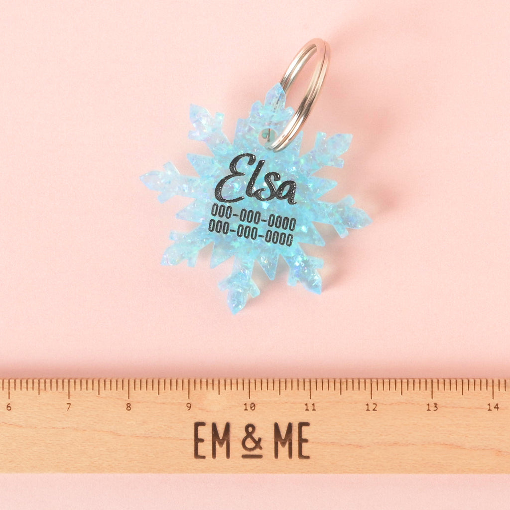 Light blue glitter snowflake-shaped pet ID tag engraved with the name Elsa and phone numbers, shown on a pink background above a wooden ruler for size reference.