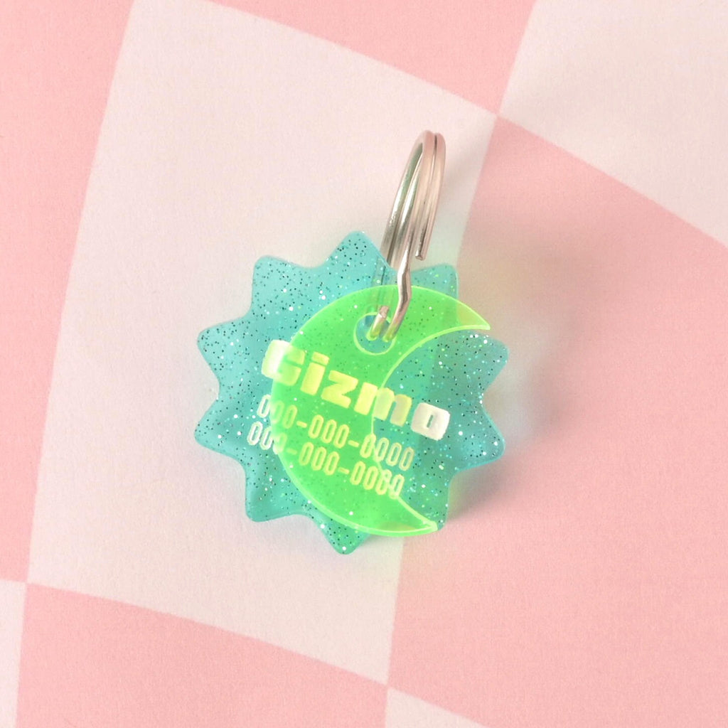 A custom, turquoise, starburst-shaped pet ID tag with a subtle glitter finish. The tag is engraved with the name "Gizmo" and a phone number. A second, translucent green crescent moon charm is attached, sitting in front of the main tag. Both are on a shiny silver keyring and are displayed on a pink and white checkered background.