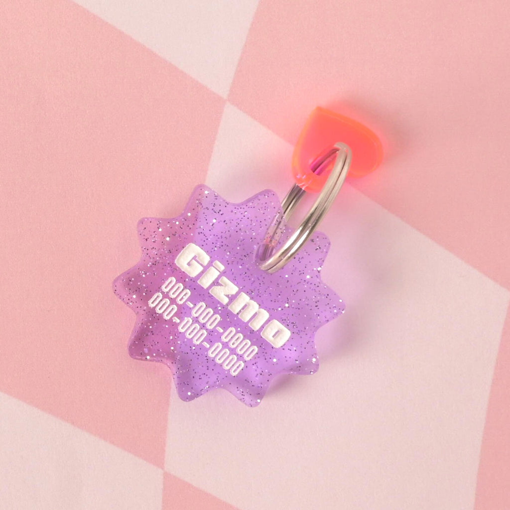 A custom, purple, starburst-shaped pet ID tag with a subtle glitter finish. The tag is engraved with the name "Gizmo" and a phone number in white font. A small Fluorescent pink heart charm is attached, sitting on top of the keyring. The tag is displayed on a pink and white checkered background.