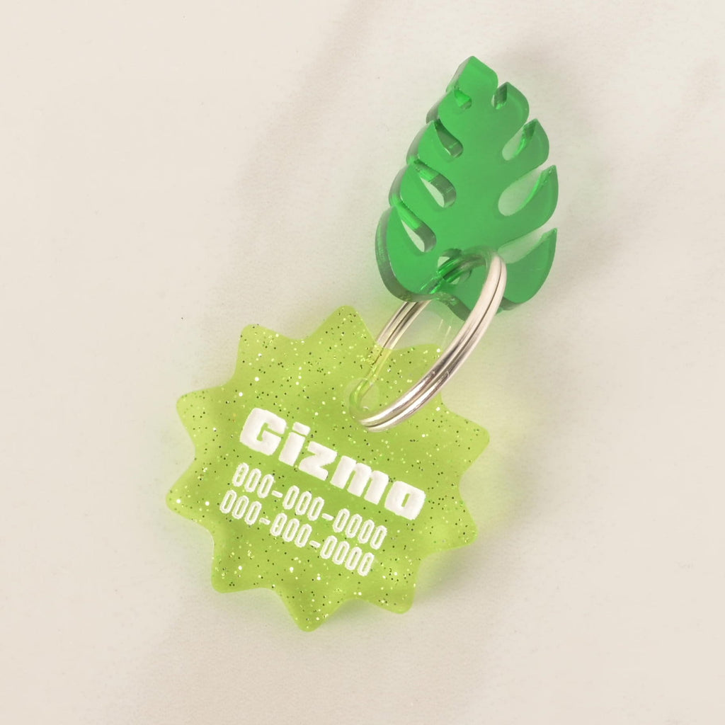 A close-up of a custom, green, starburst-shaped pet ID tag with a subtle glitter finish. The tag is engraved with the name "Gizmo" and a phone number in white font. A bright green tropical leaf charm is attached, sitting on top of the keyring. The tag is displayed on a solid white background.