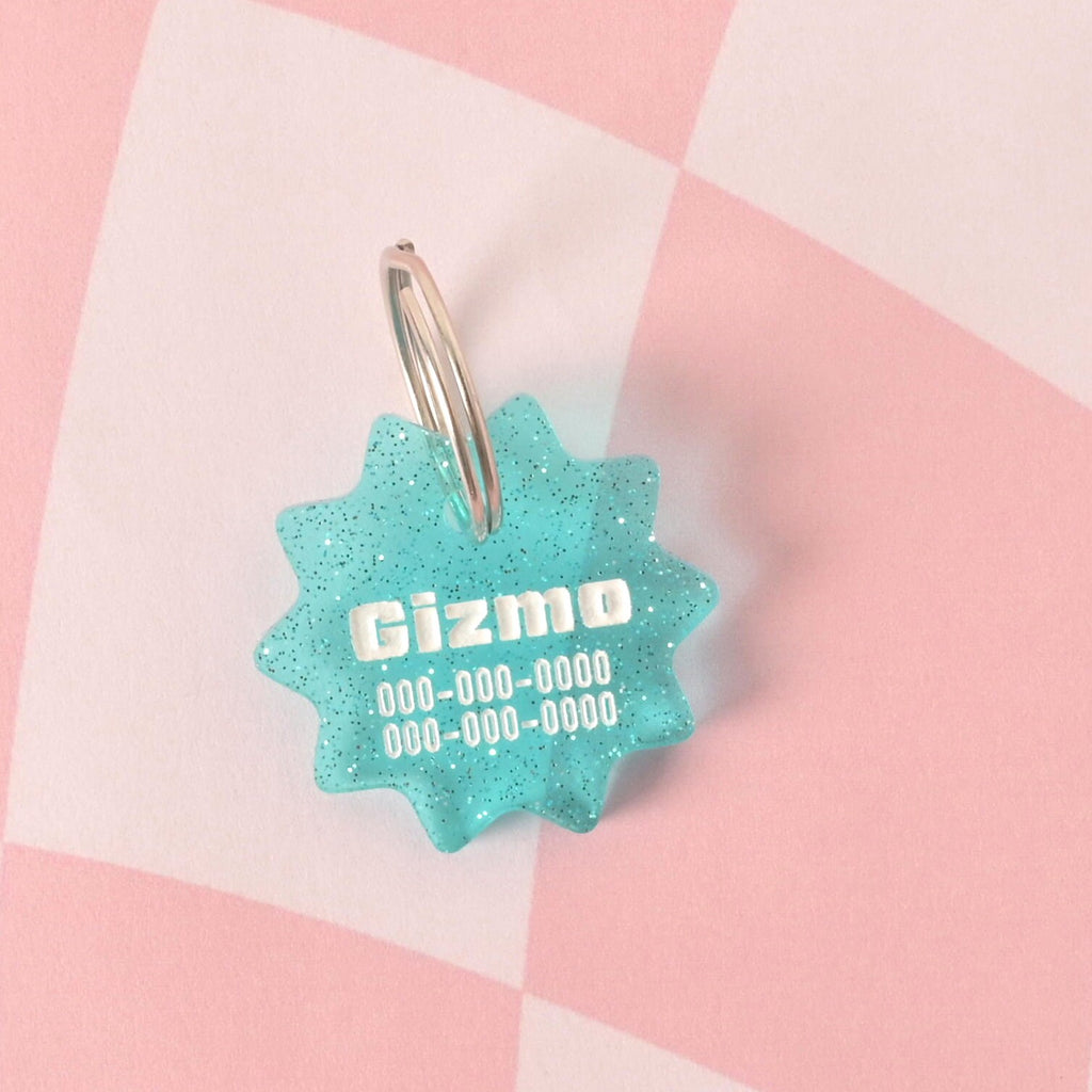 A close-up of a custom, turquoise, starburst-shaped pet ID tag. The tag has a subtle glitter finish and is engraved with the name "Gizmo" and a phone number in white font. It is attached to a shiny silver keyring and is displayed on a pink and white checkered background.