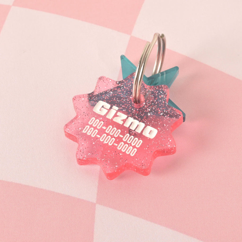 A custom, pink, starburst-shaped pet ID tag with a subtle glitter finish. The tag is engraved with the name "Gizmo" and a phone number in white font. A second translucent turquoise star tag is visible behind it. Both are attached to a shiny silver keyring and are displayed on a pink and white checkered background.
