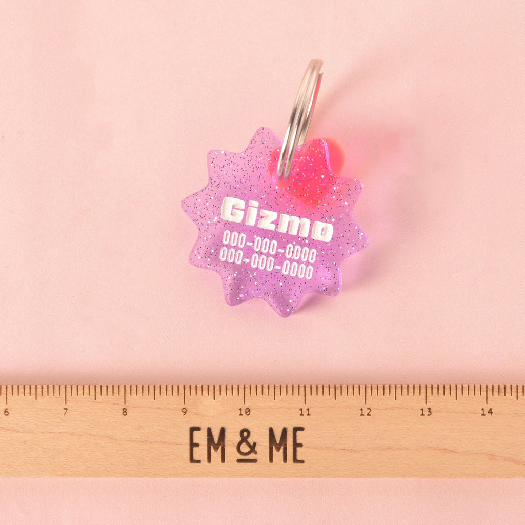 A custom, purple, starburst-shaped pet ID tag with a subtle glitter finish. The tag is engraved with the name "Gizmo" and a phone number in white font. A small translucent pink heart charm is attached, sitting on top of the keyring. A wooden ruler with the logo "EM & ME" is visible at the bottom of the photo, which is set on a pink background.