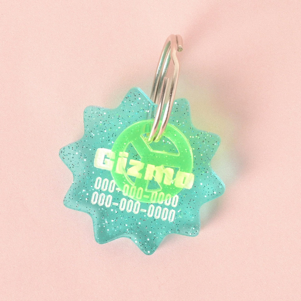 A custom, turquoise, starburst-shaped pet ID tag with a subtle glitter finish. The tag is engraved with the name "Gizmo" and a phone number in white font. A second translucent green peace sign charm is attached, sitting in front of the main tag. Both are on a shiny silver keyring and are displayed on a solid pink background.