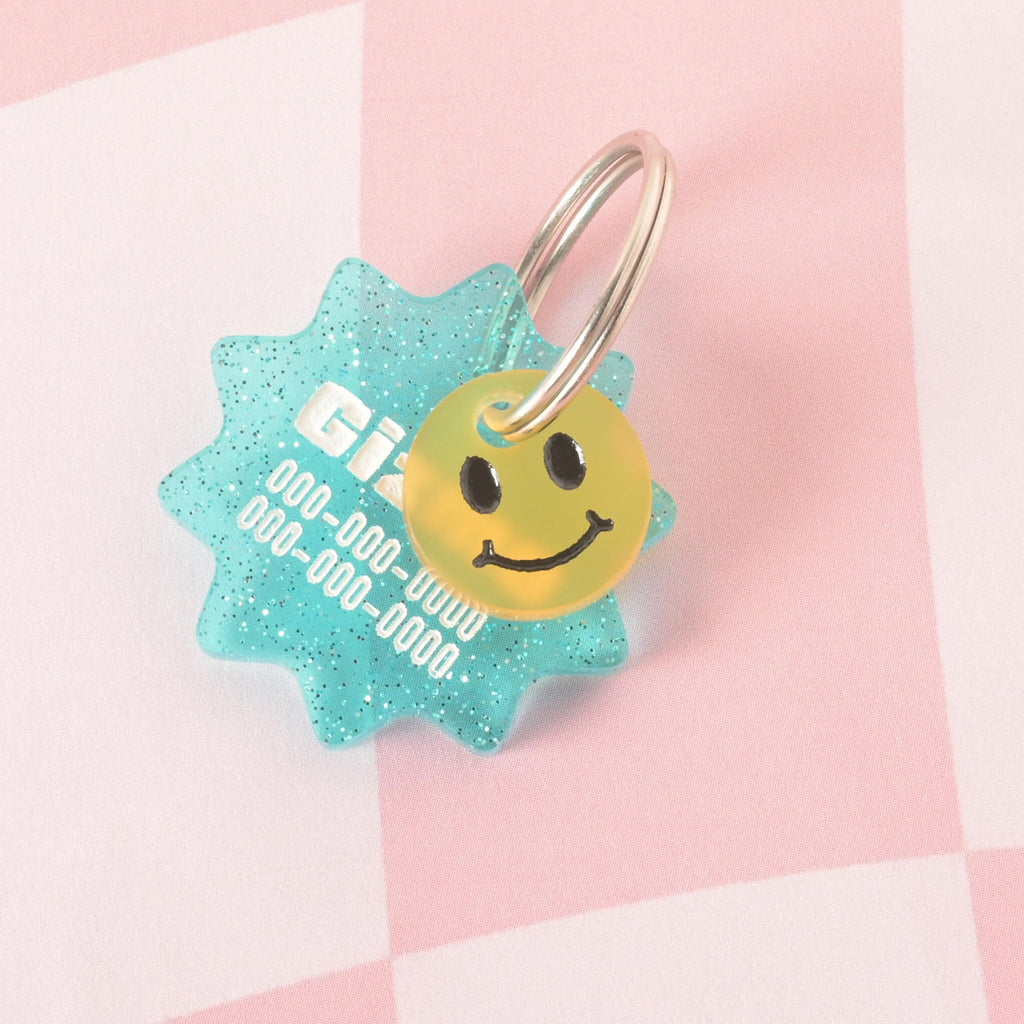 A custom, turquoise, starburst-shaped pet ID tag with a subtle glitter finish. The tag is engraved with the name "Gizmo" and a phone number in white font. A small, translucent yellow smiley face charm is attached, sitting in front of the main tag. Both are on a shiny silver keyring and are displayed on a pink and white checkered background.