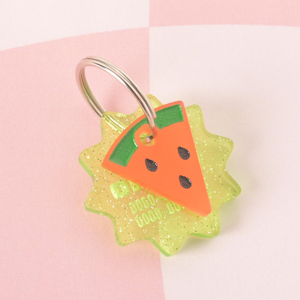 A custom, glittery yellow-green, starburst-shaped pet ID tag with a subtle phone number engraved on it. A second, pink and green watermelon slice charm with black seeds is attached to a shiny silver keyring, sitting in front of the main tag. The tags are displayed on a pink and white checkered background.