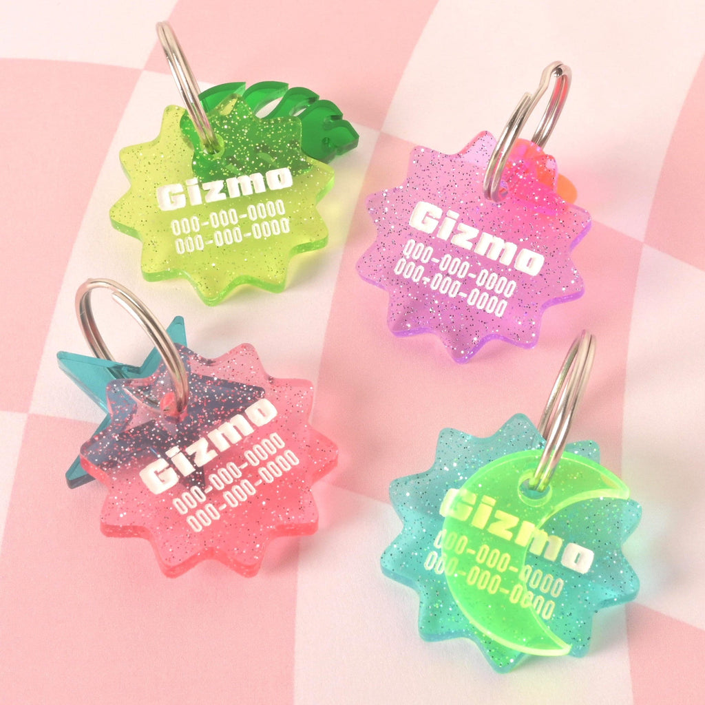 A group of four custom, starburst-shaped pet ID tags with a subtle glitter finish. The tags are in various colors: lime green with a green leaf charm, pink, light purple, and blue-green. Each tag is engraved with the name "Gizmo" and a phone number. They are attached to silver keyrings and are displayed on a pink and white checkered background.