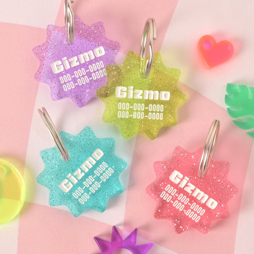 A group of four custom, starburst-shaped pet ID tags in different colors: glittery purple, green, blue, and pink. Each tag is engraved with the name "Gizmo" and a phone number. A neon orange heart charm and green leaf charm are also visible in the background. The tags are attached to silver keyrings and are displayed on a pink and white checkered background.